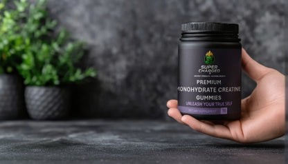 Hand holding a container of 'Charged' creatine gummies with a blurred plant and dark background